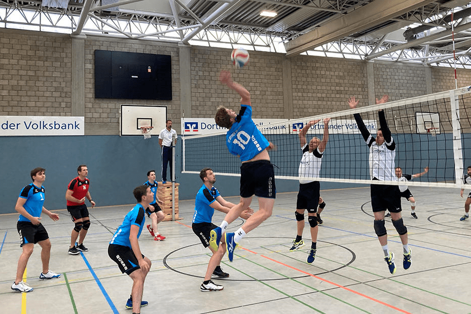 Volleyballspiel in einer Halle: Ein Spieler springt hoch, um einen Ball über das Netz zu schlagen, während andere Spieler reagieren.