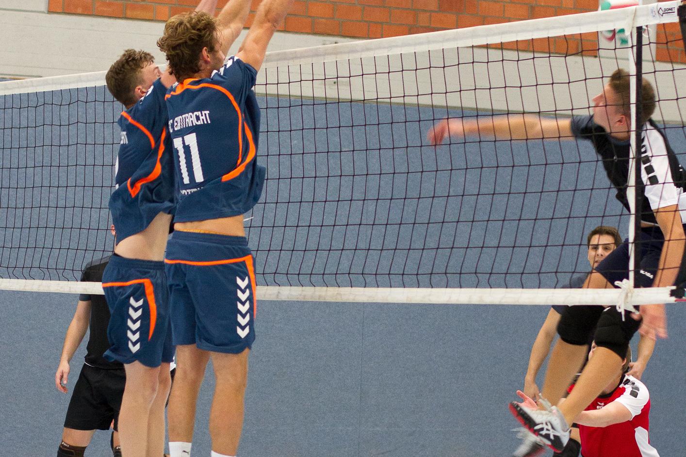 Zwei Volleyballspieler springen, um einen Ball über das Netz zu blocken, während ein dritter Spieler angreift.