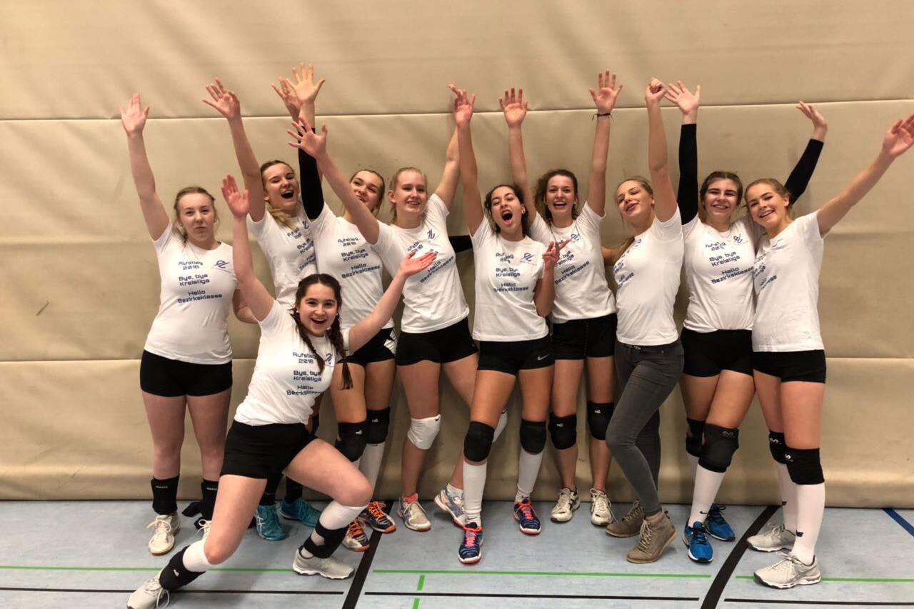 Gruppenfoto von zehn Volleyballspielerinnen in weißen T-Shirts, die fröhlich vor einer Sportwand posieren.