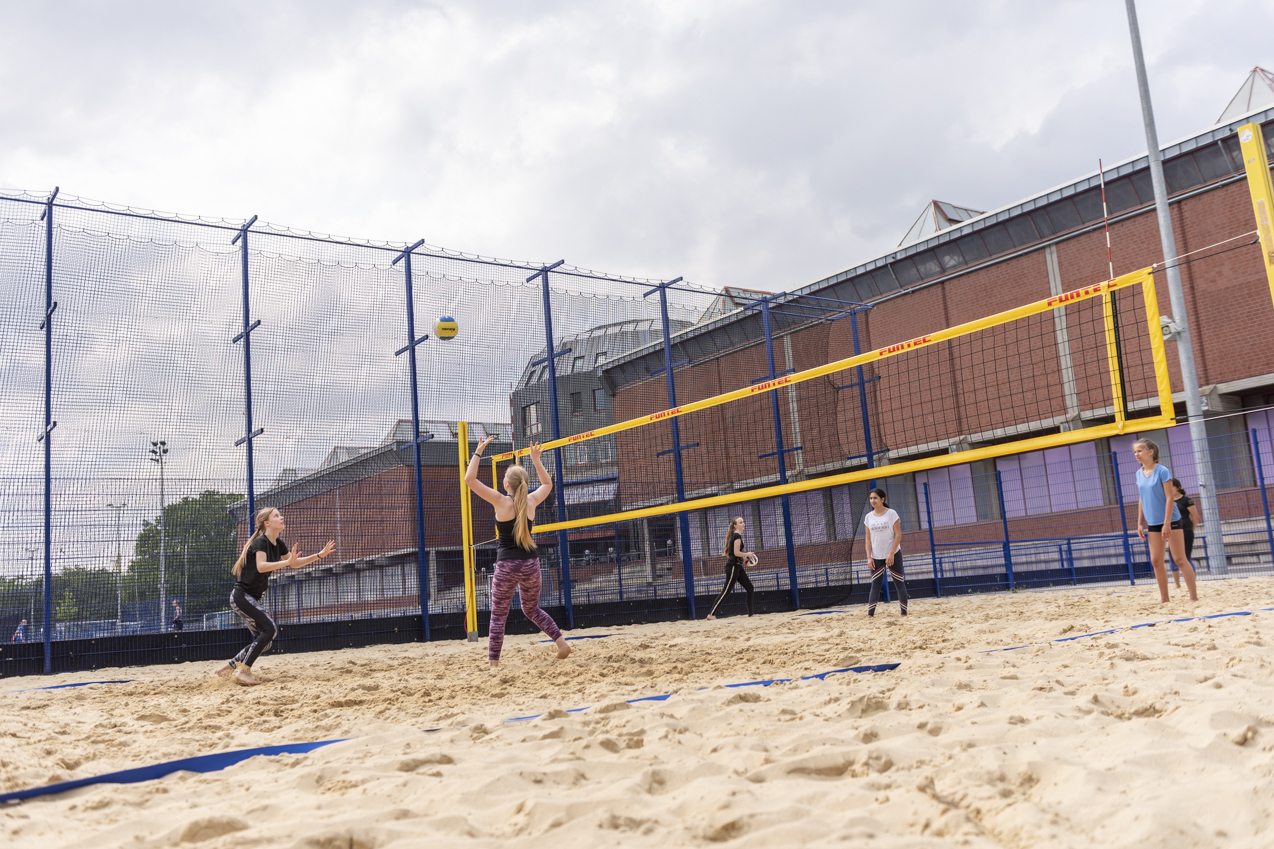 Strandvolleyballspiel mit mehreren Spielerinnen auf einem Sandplatz unter einem bew&ouml;lkten Himmel.