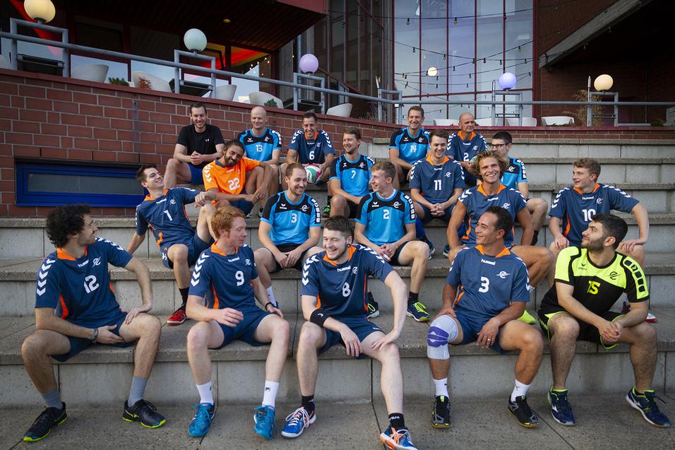 Gruppe von 15 Handballspielern in blauen Trikots, sitzend auf Treppenstufen, lächelnd und in lockerer Pose.