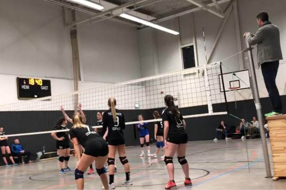 Volleyballspiel in einer Halle mit f&uuml;nf Spielerinnen in Schwarz und einer Person an der Seitenlinie mit Stoppuhr.