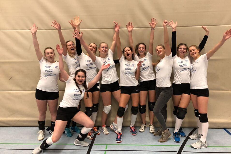 Gruppenfoto von zehn Volleyballspielerinnen in weißen T-Shirts, die fröhlich vor einer Sportwand posieren.