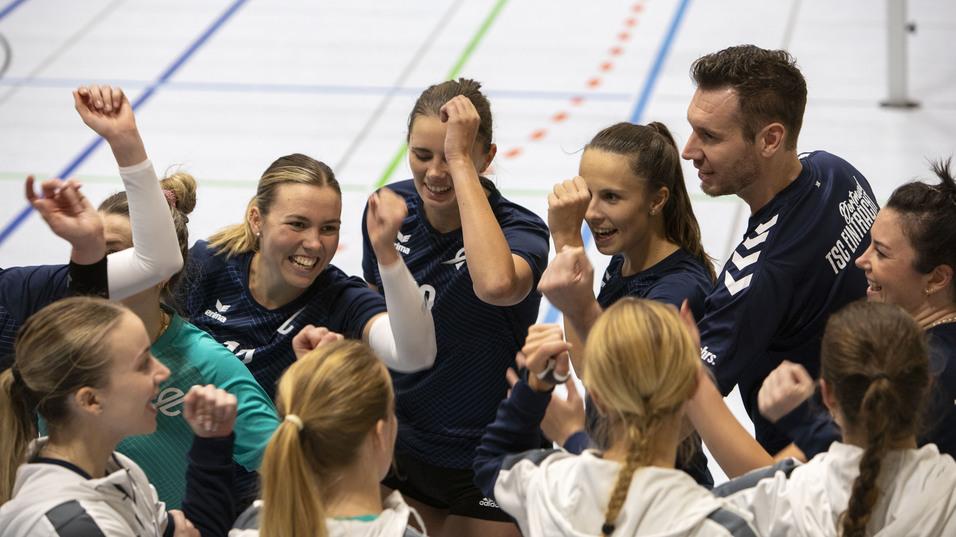 Sportteam feiert im Kreis, Spielerinnen und Trainer heben die Arme, l&auml;cheln und zeigen Teamgeist in einer Sporthalle.