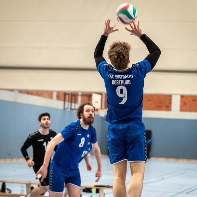 Ein Volleyballspieler hebt den Ball in die Luft, w&auml;hrend zwei Teamkollegen in einem Sporthallen-Setting zuschauen.
