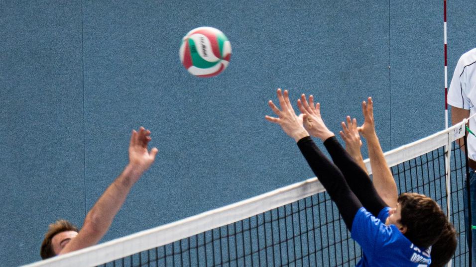 Zwei Volleyballspieler springen zum Ball &uuml;ber das Netz, w&auml;hrend ein Schiedsrichter auf einer erh&ouml;hten Plattform beobachtet.