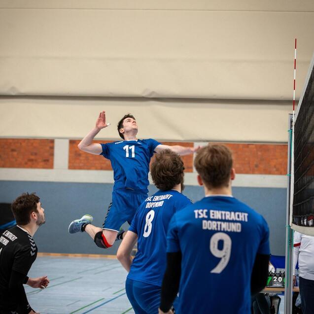 Volleyballspieler springt zum Aufschlag, w&auml;hrend Teamkollegen in blauen Trikots auf dem Spielfeld stehen.