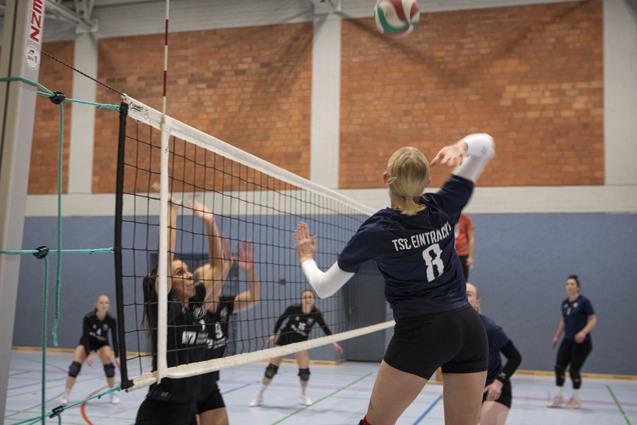 Spielszene eines Volleyballspiels, mit einer Spielerin, die den Ball &uuml;bers Netz schl&auml;gt, w&auml;hrend Gegnerinnen reagieren.