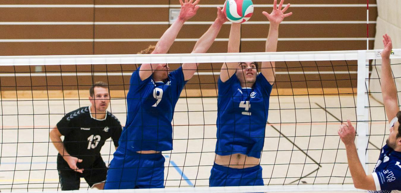 Zwei Volleyballspieler in blauen Trikots blockieren mit erhobenen Armen einen auf sie zuschnellenden Volleyball.