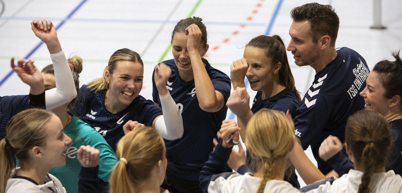 Sportteam feiert im Kreis, Spielerinnen und Trainer heben die Arme, l&auml;cheln und zeigen Teamgeist in einer Sporthalle.