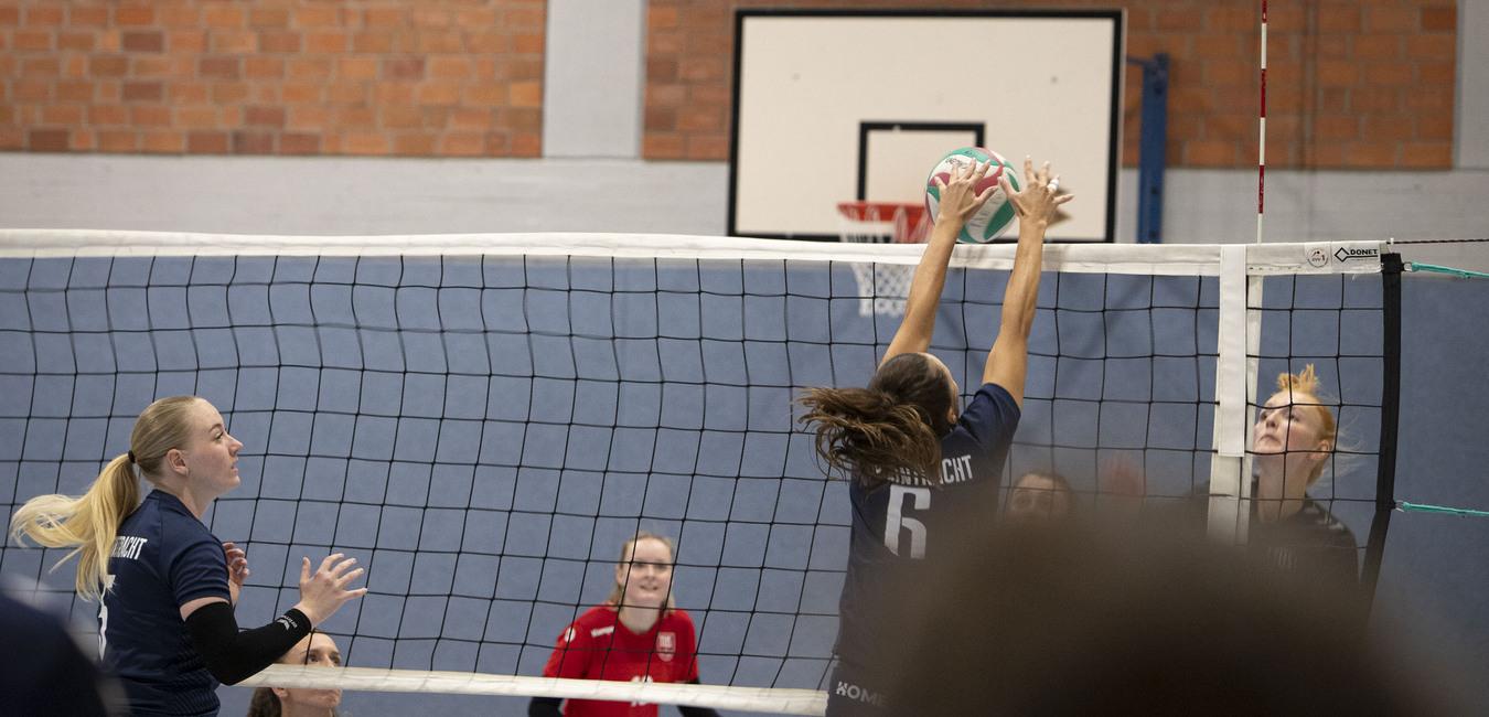 Spielszene im Volleyball: Spielerinnen springen, um den Ball &uuml;ber das Netz zu schlagen, w&auml;hrend andere Zuschauer beobachten.