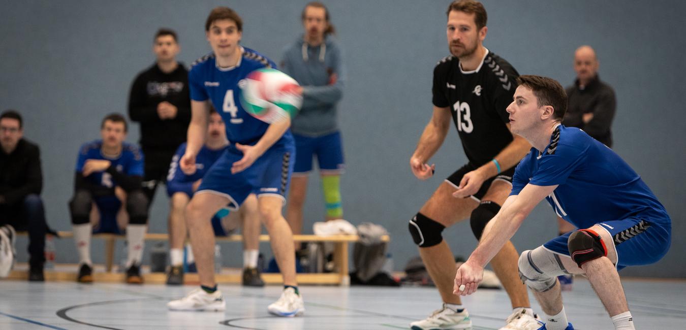 Zwei Volleyballspieler in blauen Trikots bereiten sich auf den Ballkontakt in einer Sporthalle vor, Zuschauer im Hintergrund.