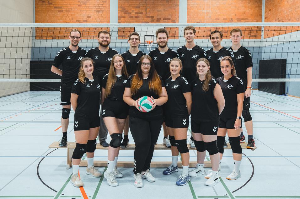 Gruppenfoto eines Volleyballteams in Sportkleidung, das in einer Halle vor einem Volleyballnetz posiert.