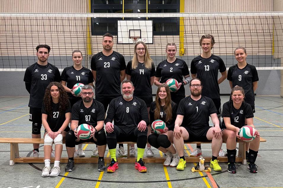 Gruppenfoto einer Volleyballmannschaft in schwarz-wei&szlig;en Trikots, posed vor einem Netz in einer Halle.