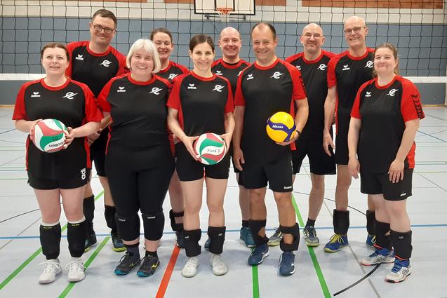 Gruppe von zehn Volleyballspielern in schwarz-roten Trikots, posierend auf einem Sportfeld mit Bällen in der Hand.