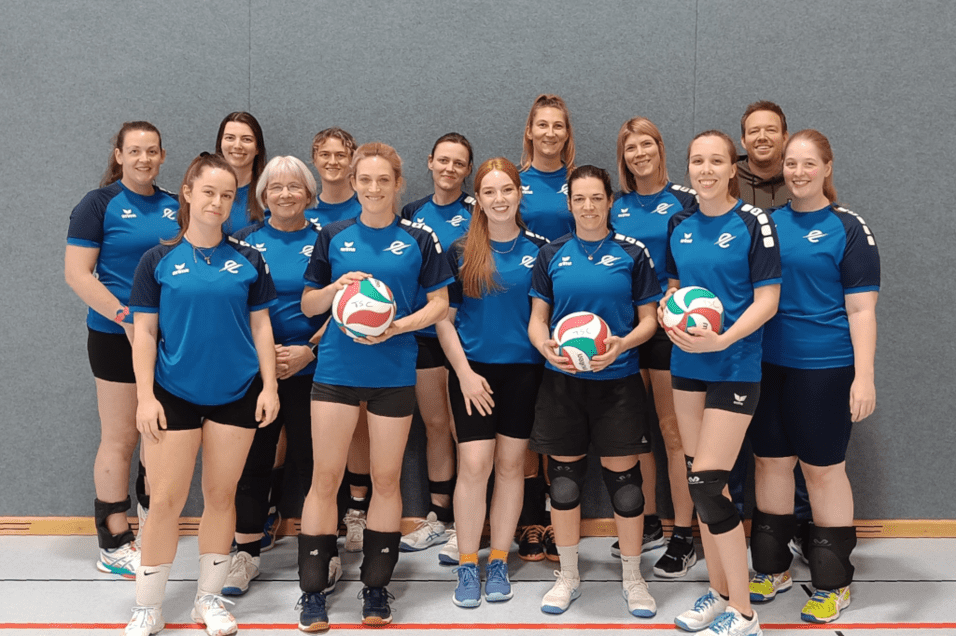 Gruppenfoto eines Volleyballteams in blauen Shirts, alle halten einen Volleyball und posieren vor grauer Wand.