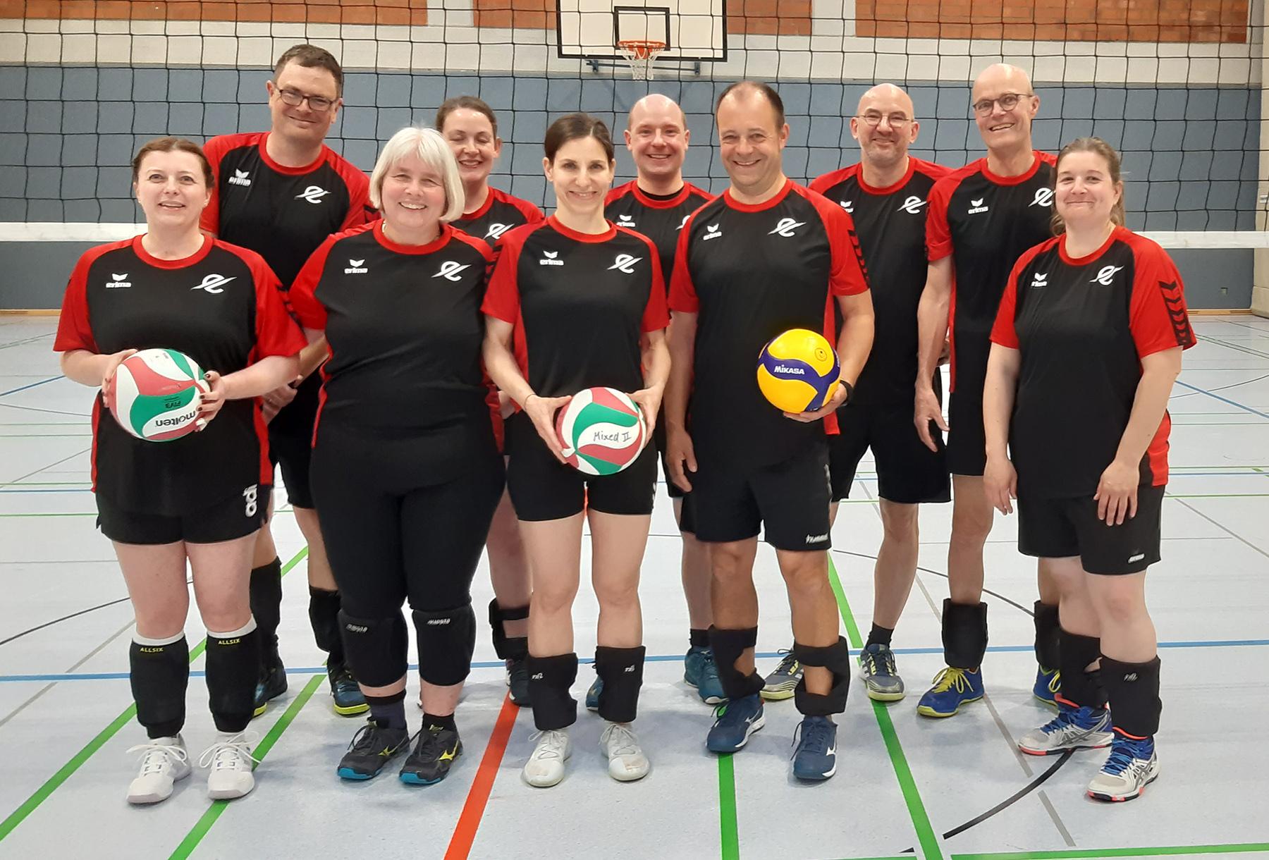 Gruppe von zehn Volleyballspielern in schwarz-roten Trikots, posierend auf einem Sportfeld mit B&auml;llen in der Hand.