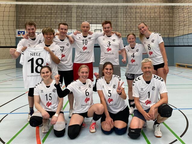 Gruppe von zehn Volleyballspielern in weißen Trikots, posierend auf einem Volleyballfeld vor dem Netz.