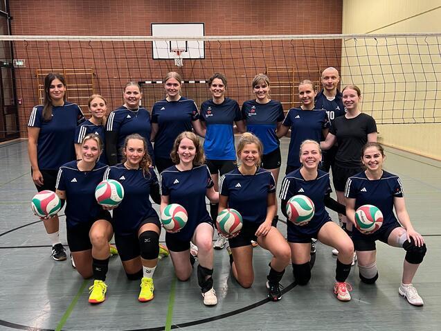 Gruppenfoto einer Volleyballmannschaft mit 16 Frauen, die in Sportkleidung stehen und Volleyballs halten.