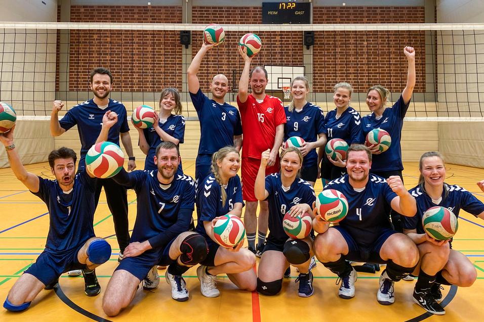Gruppe von Volleyballspielern in blauen Trikots, die fr&ouml;hlich mit B&auml;llen vor einem Netz posieren.