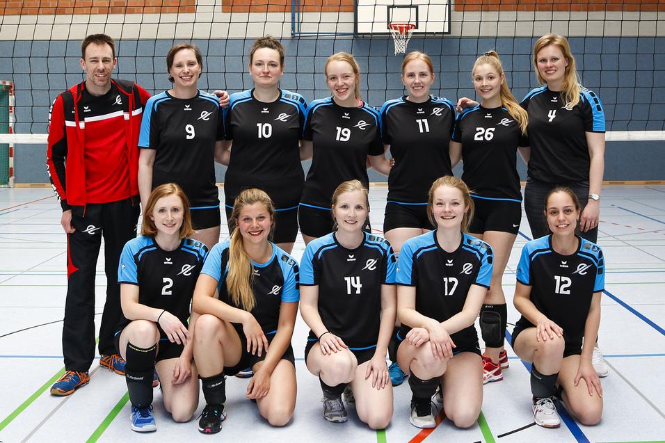 Mannschaftsfoto einer Volleyballmannschaft mit Trainer in Sportkleidung vor einem Volleyballnetz in einer Halle.