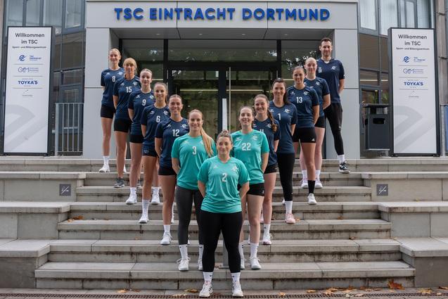 Gruppenfoto des Volleyballteams TSC Eintracht Dortmund vor dem Vereinsgebäude, Spielerinnen in blau und grün, lächelnd.