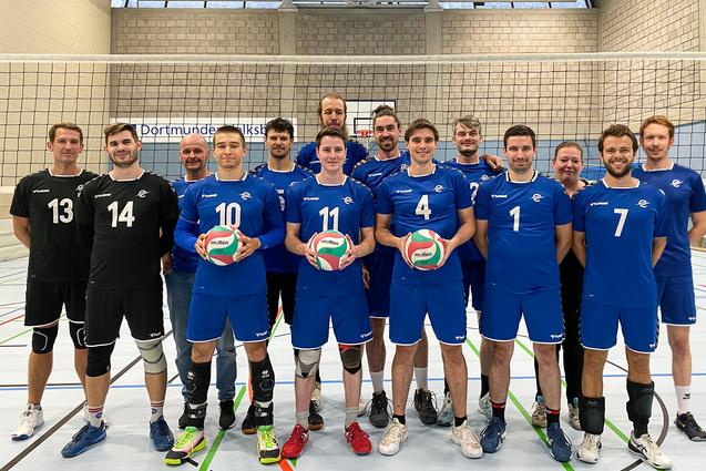Gruppenzusammenstellung von Volleyballspielern in blauer Teamkleidung, vor einem Volleyballnetz, mit Bällen in den Händen.