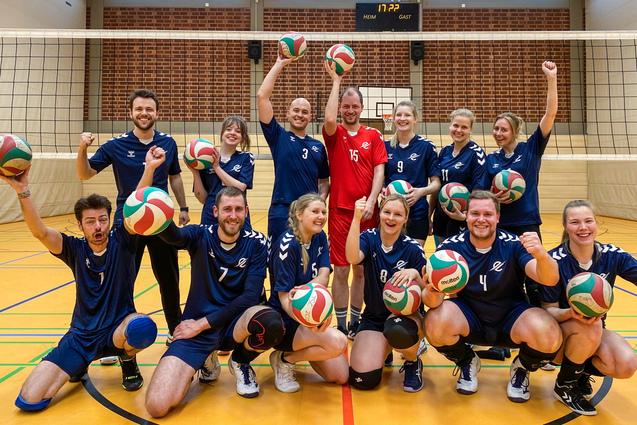 Mixed 6 Gruppe von Volleyballspielern in blauen Trikots, die fröhlich mit Bällen vor einem Netz posieren.
