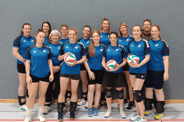 Gruppenfoto eines Volleyballteams in blauen Shirts, alle halten einen Volleyball und posieren vor grauer Wand.