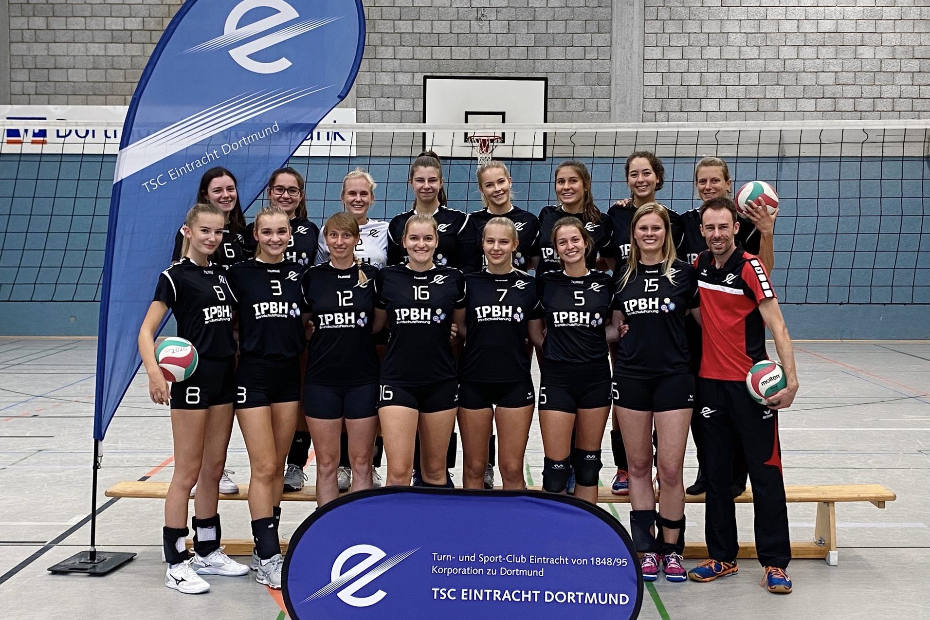 Gruppennfoto des weiblichen Volleyballteams TSC Eintracht Dortmund vor einem Netz, mit Bällen und Banner im Hintergrund.