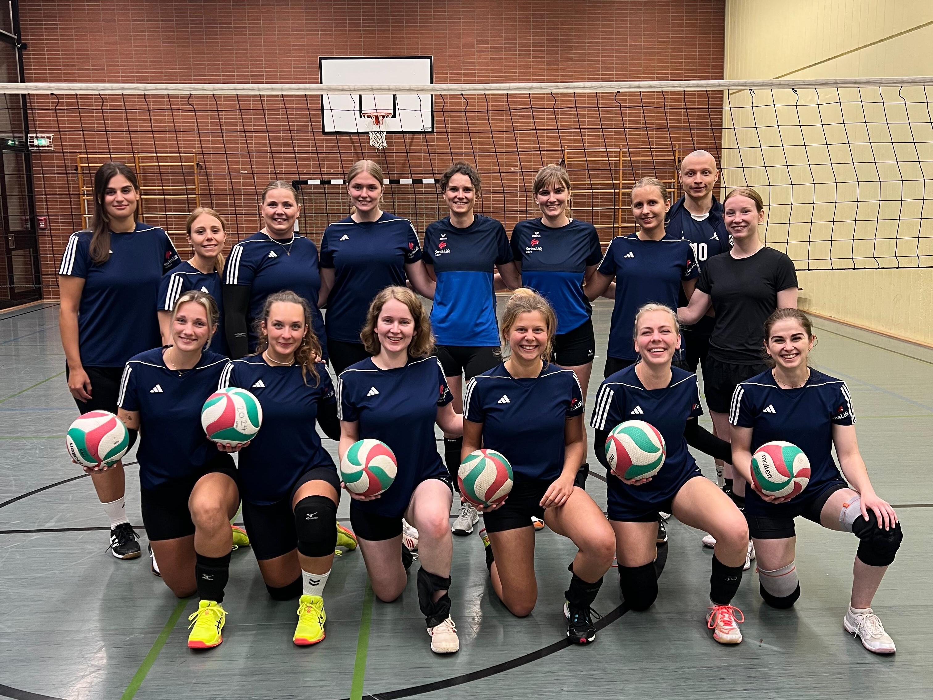 Gruppenfoto einer Volleyballmannschaft mit 16 Frauen, die in Sportkleidung stehen und Volleyballs halten.