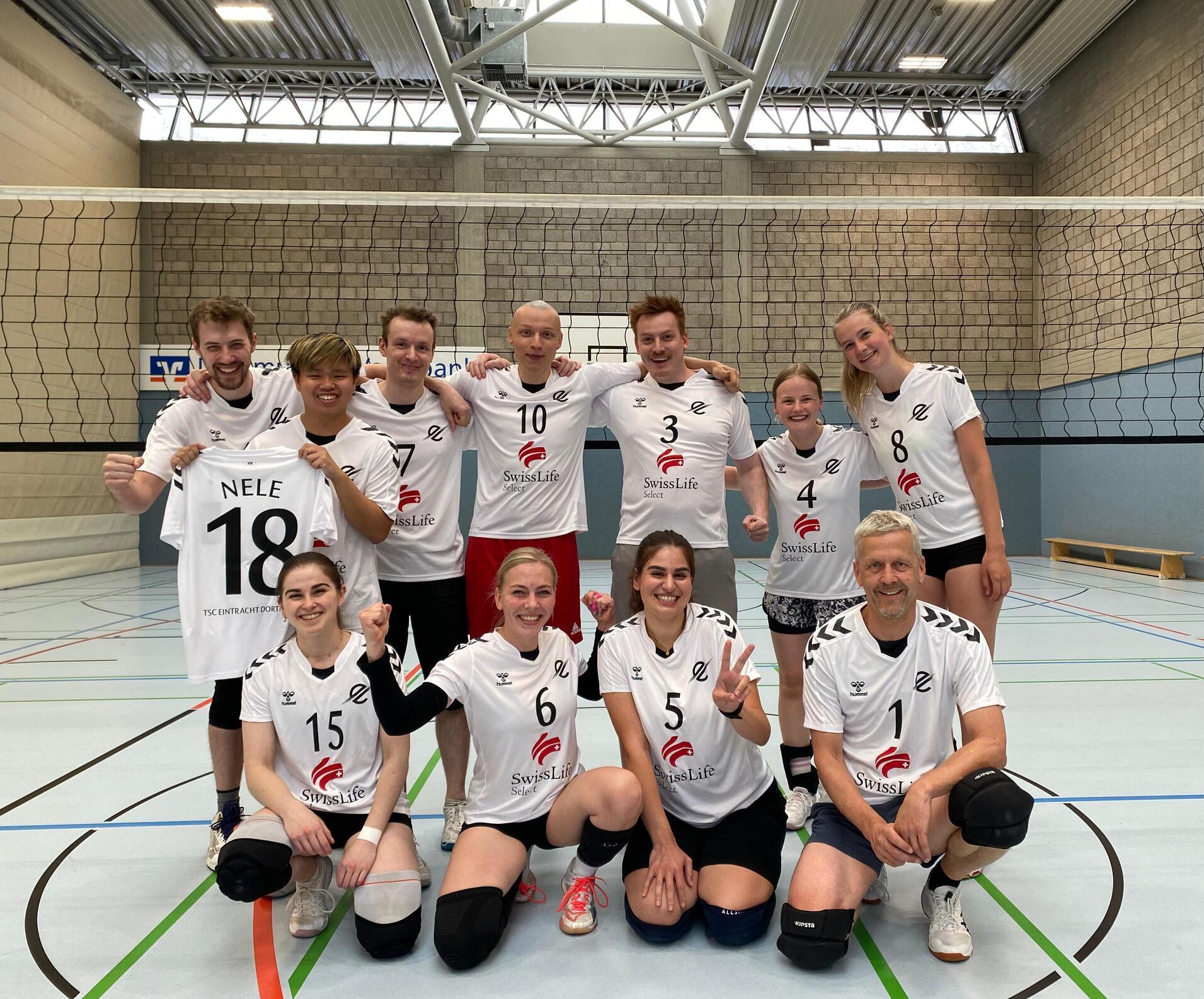 Gruppe von zehn Volleyballspielern in weißen Trikots, posierend auf einem Volleyballfeld vor dem Netz.