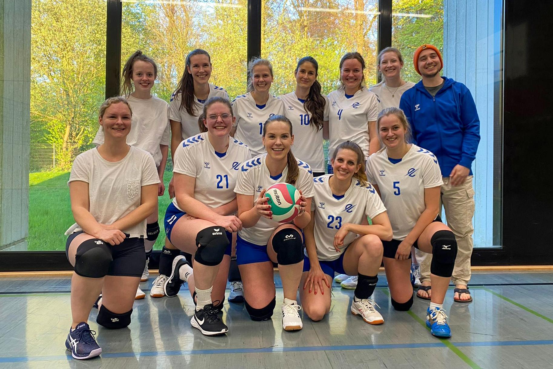 Gruppennahaufnahme eines Volleyballteams mit 12 Spielerinnen und einem Trainer, alle in sportlichen Outfits, lächeln.