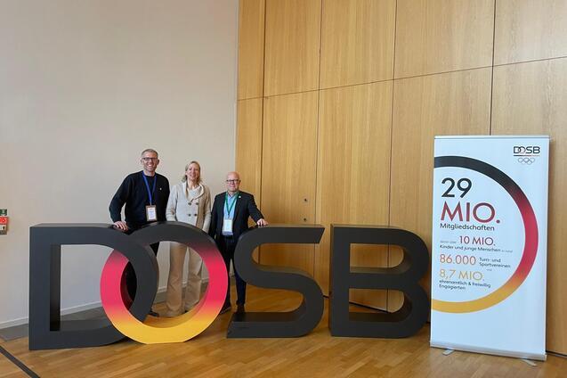 Drei Personen stehen neben einer gro&szlig;en, bunten &bdquo;DOSB&ldquo;-Buchstaben-Darstellung vor einer Wand. Rechts ein Informationsbanner.