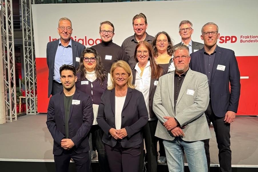 Gruppenfoto von zw&ouml;lf Personen auf einer B&uuml;hne, alle l&auml;chelnd vor einem Hintergrund mit SPD-Logo.