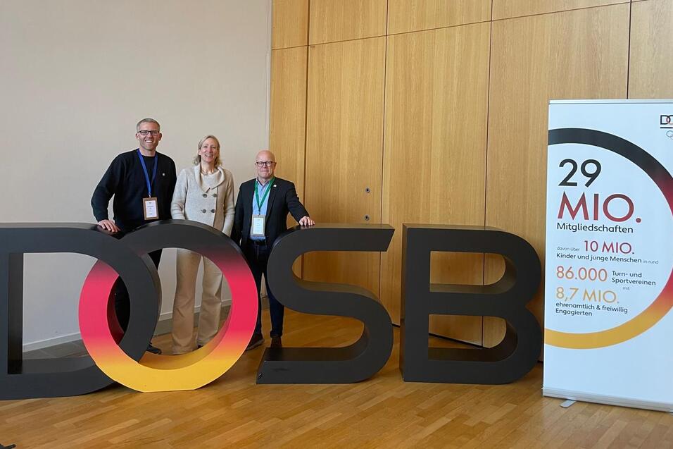 Drei Personen stehen neben einer gro&szlig;en, bunten &bdquo;DOSB&ldquo;-Buchstaben-Darstellung vor einer Wand. Rechts ein Informationsbanner.