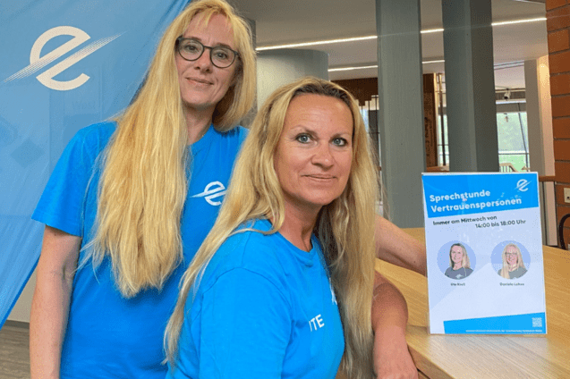 Zwei Frauen in blauen T-Shirts stehen an einem Tisch, neben einer Informationsbrosch&uuml;re und einem blauem Fahne.