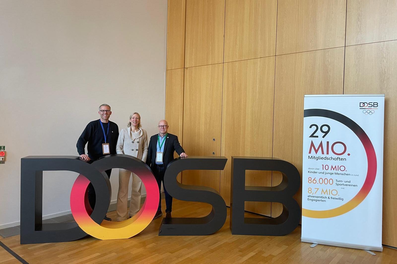 Drei Personen stehen neben einer großen, bunten „DOSB“-Buchstaben-Darstellung vor einer Wand. Rechts ein Informationsbanner.