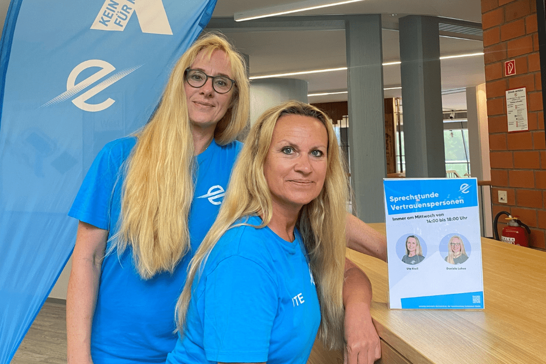 Zwei Frauen in blauen T-Shirts stehen an einem Tisch, neben einer Informationsbroschüre und einem blauem Fahne.