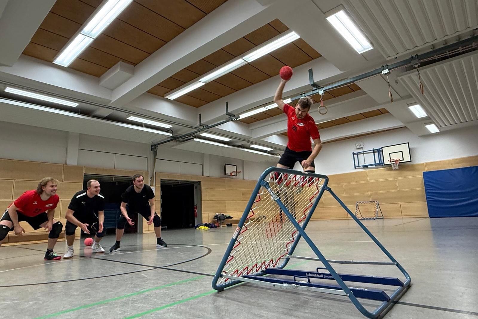 Spieler in rotem Trikot werfen einen Handball auf ein Tor, während drei Teamkollegen bereit stehen.