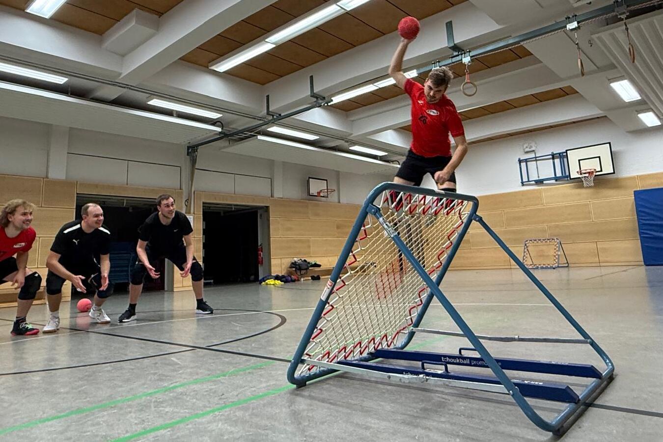 Spieler in rotem Trikot werfen einen Handball auf ein Tor, w&auml;hrend drei Teamkollegen bereit stehen.