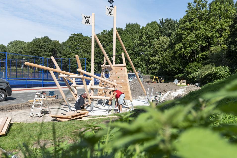 Zwei Arbeiter montieren eine Holzspielplatzstruktur mit Kletterwand und drehbaren Elementen in einem Park.