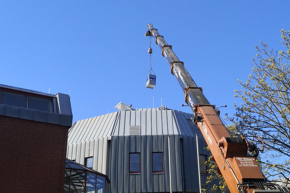 Kran hebt eine Arbeitsbühne an das Dach eines modernen Gebäudes unter blauen Himmel.
