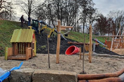 Baustelle eines Spielplatzes mit Holzspielhaus, Rutsche und Baggerarbeit im Hintergrund. Sandfläche und Sicherheitsbereich sichtbar.