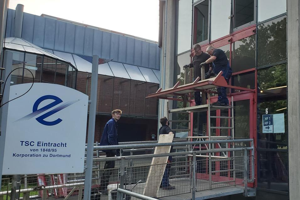 Arbeiter stehen auf einem Gerüst, während sie ein Fenster an einem modernen Gebäude renovieren. Ein TSC Eintracht-Schild ist im Vordergrund sichtbar.