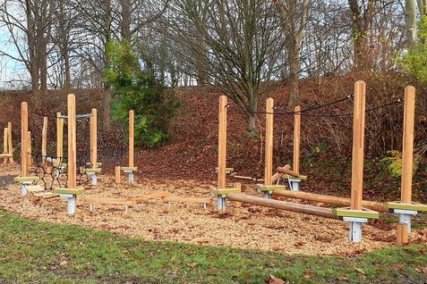 Outdoor-Fitnessbereich mit Holzgeräten und einem geschotterten Untergrund, umgeben von Bäumen und Sträuchern.