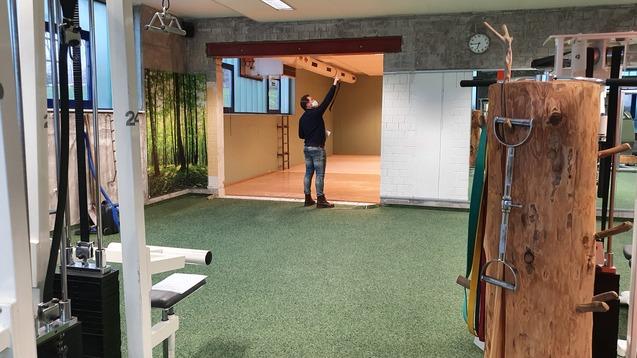 Mann mit Arbeitshelm prüft die Decke eines Fitnessraums mit Teppichboden und Holzelementen. Fitnessgeräte im Vordergrund.