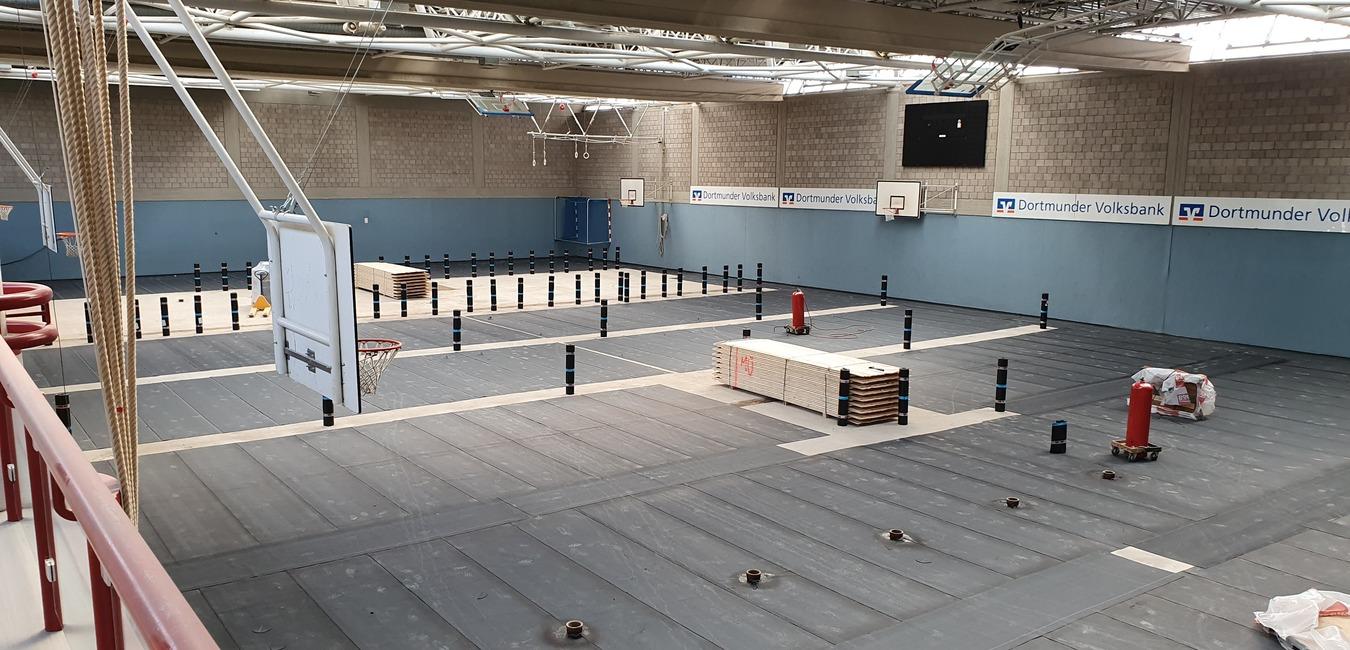 Blick in eine große Sporthalle mit Holzständen, Basketballkörben und mehreren Markierungsstangen auf dem Boden.