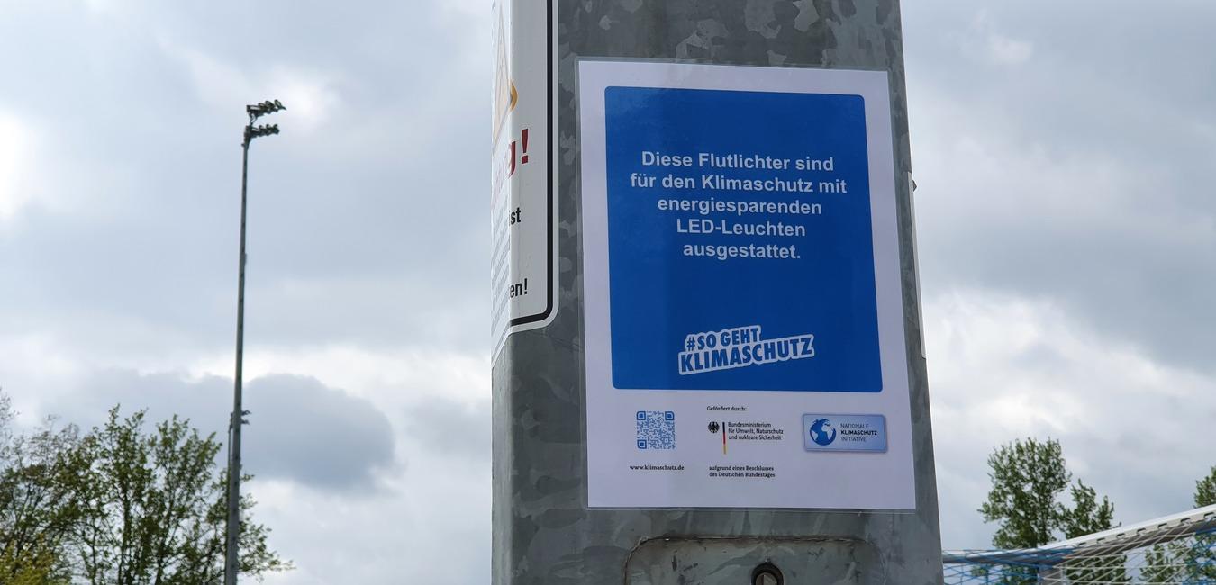 Hinweis auf einem Mast: "Flutlichtanlagen f&uuml;r Klimaschutz mit energiesparenden LED-Leuchten ausgestattet."