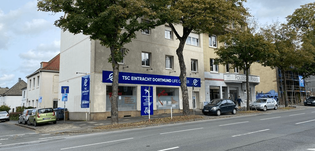 Mehrst&ouml;ckiges Geb&auml;ude mit TSC Eintracht Dortmund-Werbung, umgeben von B&auml;umen und parken Autos auf einer Stra&szlig;e.